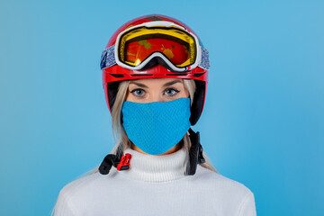Close-up portrait of a girl skier wearing a light blu protective mask, helmet and glasses. A snowboarder on an light blu background. Covid19 coronavirus and the ski resort pandemic.