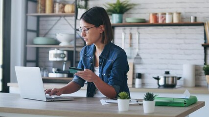 Video of cute young woman holding white credit card for shopping online with computer while sitting in the kitchen at home. - Powered by Adobe