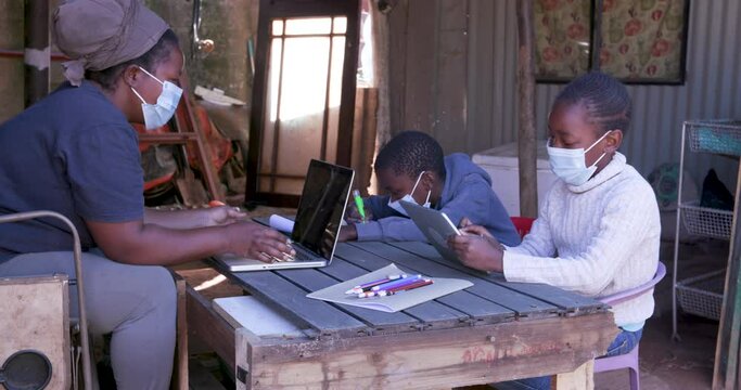 Black African woman using a laptop and digital tablet to home school her two young children at their dilapidated home during lockdown for Covid-19 Coronavirus pandemic, South Africa