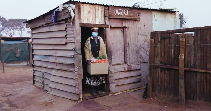 Close-up of an old Black African woman standing in the door way of her tin shack in an informal settlement with a food parcel donated to her by an NGO during Covid-19