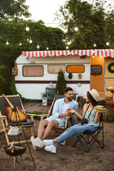 Happy couple with burgers and beer resting near trailer. Camping season - obrazy, fototapety, plakaty