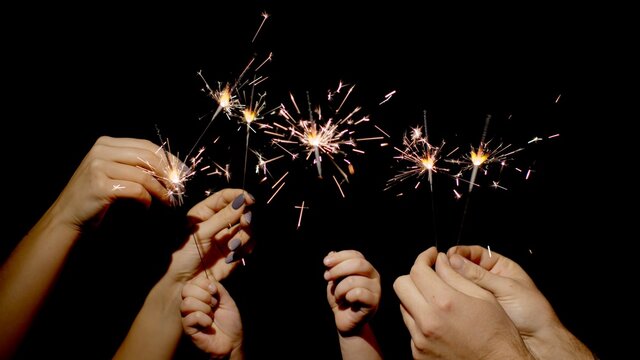 Close-up of hands or palms holding and waving burning sparklers in front of black dark background. Sparkling lights at birthday party, wedding, New Year, Christmas Eve, Xmas. 6k downscale. Bengal fire