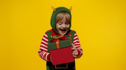 Joyful kid girl in Christmas elf Santa helper costume on yellow background. Child receiving present gift box, expressing amazement, extreme happiness looking inside box. New Year holidays celebration