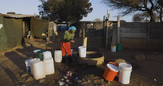 Face of poverty.  Poor Black African woman with no access to running water, drinks water while filling up buckets at a central tap to take to her house in an informal settlement (squatter camp) 