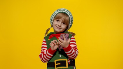 Little kid child girl in Christmas elf Santa helper costume types something on mobile phone display enjoys browsing social media sends messages of congratulations, online gifts. Yellow background