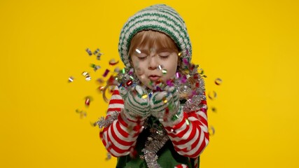 Kid girl in Christmas elf Santa helper costume having fun blows confetti to the camera isolated on yellow background. Child blowing confetti, fooling around. People New Year holidays celebration