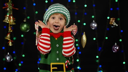 Shy shamed child girl in Christmas elf Santa helper costume posing and making funny faces. Kid...