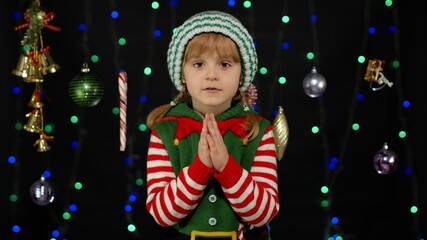 Fototapeta na wymiar Pleading kid in Christmas elf Santa helper costume begs, prays for forgiveness feels sorry and guilty, keeps palms together, standing over black background. Child girl pleads about gifts on New Year