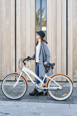 Fototapeta premium Portrait young pretty Caucasian girl, dressed in a gray jacket, a white sweater and a hat with a bike ride in the city