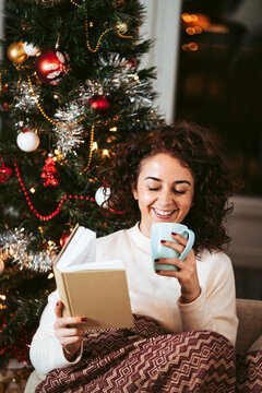 A beautiful woman laughing while reading a book in a cold day on Christmas. She is covered with a blanket while enjoying a cup of tea. Christmas holidays concept