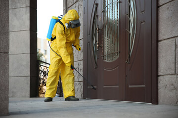 Person in hazmat suit disinfecting entrance door with sprayer. Surface treatment during coronavirus pandemic