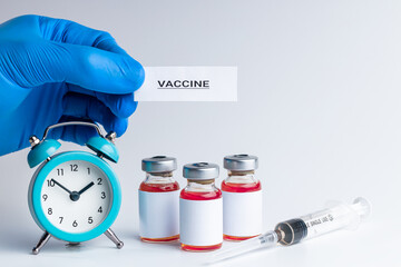 medical vaccine vials with red liquid and alarm clock over gray background. time to vaccination...