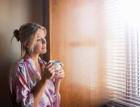 Thoughtful young woman in bathrobe looking through window while having coffee at home - Powered by Adobe