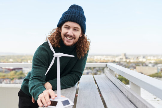 Smiling male hipster with windmill model on building terrace against sky