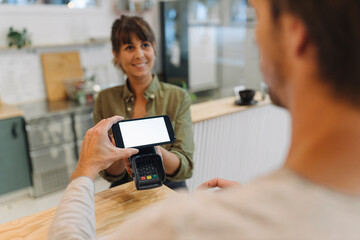 Customer doing contactless payment with smart phone to female owner at checkout in coffee shop