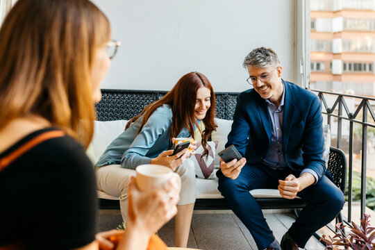 Smiling businessman showing smart phone to coworker while having coffee on sofa in balcony