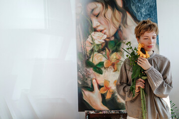 Thoughtful mid adult female artist holding sunflower while standing against artwork in studio