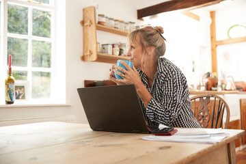 Smiling senior woman looking away while drinking coffee sitting with laptop in kitchen at home
