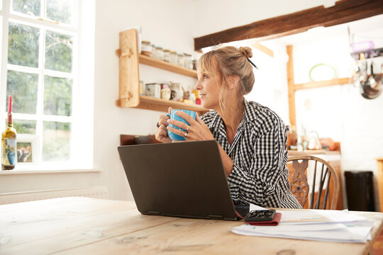 Senior woman looking away while drinking coffee sitting with laptop in kitchen at home - Powered by Adobe