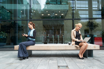 Businesswoman and colleague working while sitting with social distance on bench against office building exterior