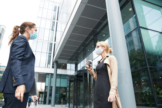 Business people wearing face mask talking while standing with social distance against office building