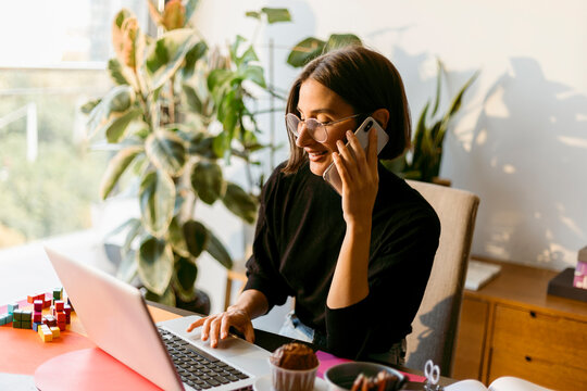 Mid adult businesswoman talking on smart phone while using laptop sitting at home