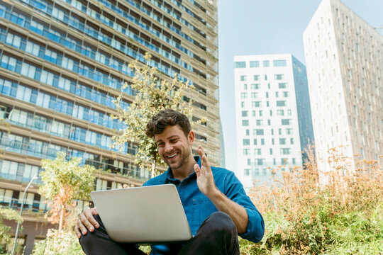 Happy young businessman waving hand while video calling through laptop against modern buildings