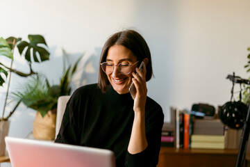 Smiling businesswoman talking on smart phone while using laptop sitting at home
