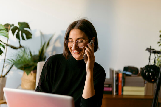 Smiling businesswoman talking on smart phone while using laptop sitting at home - Powered by Adobe