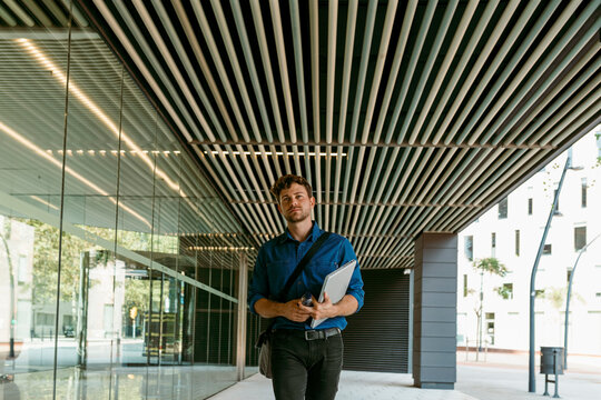 Young businessman walking with laptop on walkway at modern office building