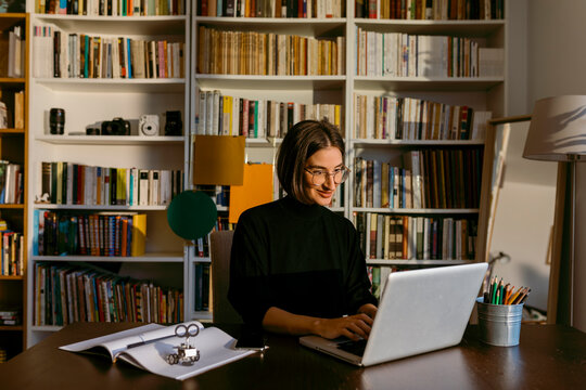 Smiling businesswoman working on laptop sitting against bookshelf at home
