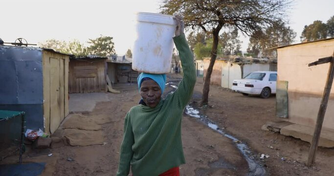 Face of poverty.  Poor Black African woman with no access to running water, walks to her house carrying a bucket of water on her head in an informal settlement (squatter camp). South Africa
