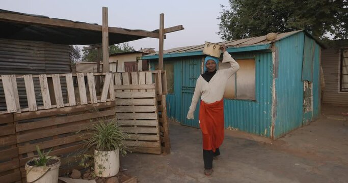 Face of poverty.  A poor Black African woman walking with a rusty tin container on her head from her densely over crowded and populated shack in an informal settlement (squatter camp). South Africa