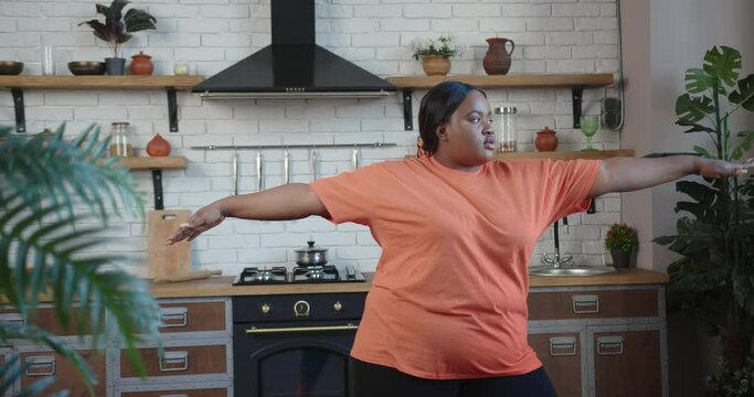 Concentrated overweight African-American dark haired woman with ponytail practices yoga asana standing in warrior pose in kitchen