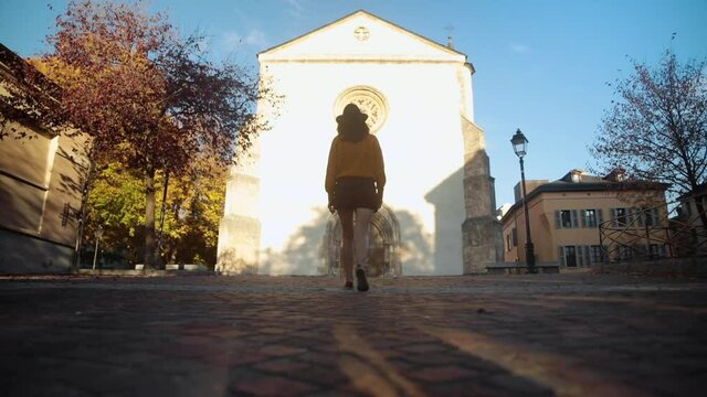 Brunette woman wearing a hat, walking towards a church in autumn sunset lights