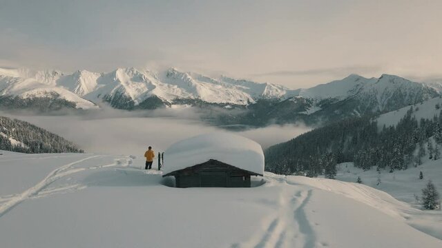Splitboarder stands next to a wooden hut with fresh snow on the roof.