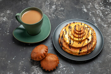 Above view of delicious pancake with two cakes on black plate with a cup of coffee on gray background