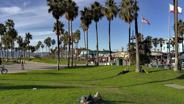 People relaxing in the grass at the Venice Beach Boardwalk enjoying the day on a sunny afternoon with the American and California flag flying in the background in Los Angeles, California, USA