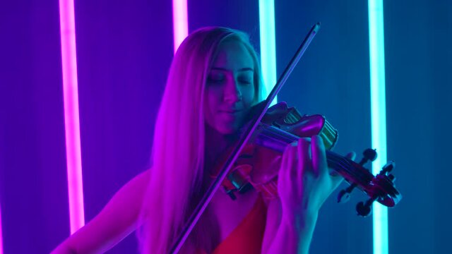 Happy smiling female artist enjoys playing the violin. A cute blonde woman performs in a dark studio against a background of bright neon lights. Close up. Slow motion.