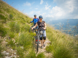 Man and woman mountain bikers walking down rough trail in beautiful landscape in Italy