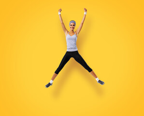 Fototapeta na wymiar Full body of happy excited woman jumping or doing fitness exercise, isolated over yellow color background. Fit girl with raised up arms, in grey sportswear, at studio shot.