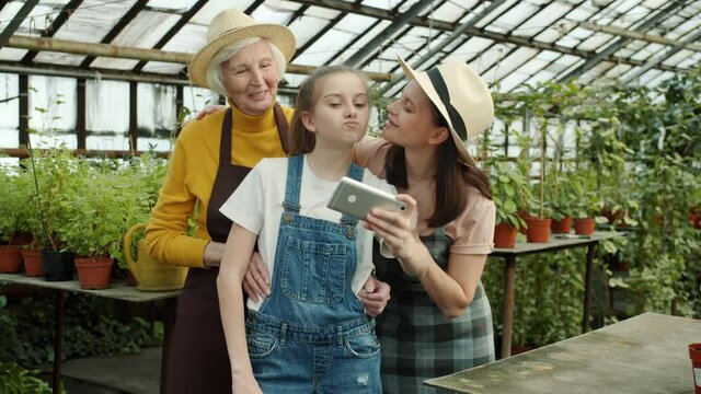 Girl is taking selfie with mother and grandmother farmers in aprons in beautiful green glasshouse using smartphone camera posing kissing smiling