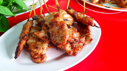 Thai grilled pork skewers in dish or plate on red table with blurred basil or thyme leaves, vegetable and other food background at local street food. Asian food and delicious meal.
