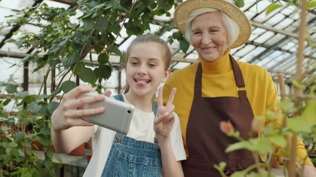 Happy girl taking selfie with loving granny posing for smartphone camera in greenhouse making funny faces smiling having fun. People and photos concept.