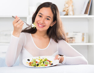 Young smiling girl eating tasty salad sitting on the table