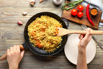Woman putting pilaf into plate from frying pan on wooden table