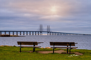 Fototapeta na wymiar Two benches in front of a beautiful ocean view with a bridge. Blue ocean and a sunset sky in the background. Picture from Malmo, southern Sweden