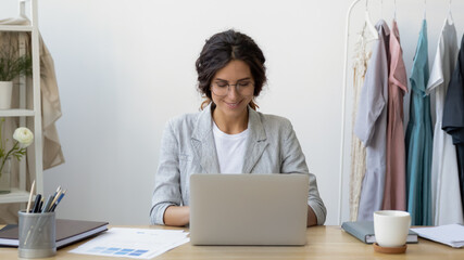 Starting workday. Happy young businesswoman self-employed tailor receive customers orders by email. Smiling lady fashion designer sitting at desk. Receptionist in atelier sewing workshop using laptop