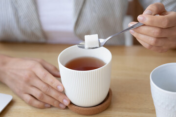 Time for tea. Close up of woman student employee taking short break in work preparing to enjoy hot drink at workplace. Young female holding lamp of sugar on teaspoon above cup of flavored tea blend