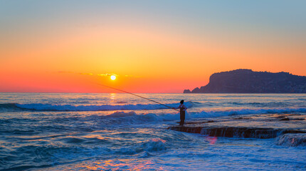 Silhouette of a fisherman on the sea embankment with rocks, Alanya castle at sunset in the background - Alanya, Turkey